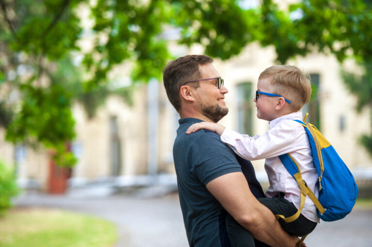Smiling man wearing glasses holding a child with glasses and backpack.