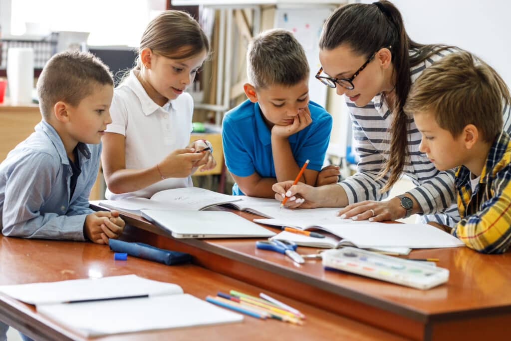 Teacher surrounded by students while writing in a notebook.