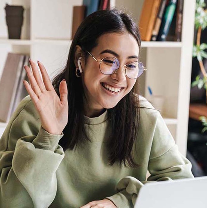 Smiling woman wearing glasses and waving at a laptop.