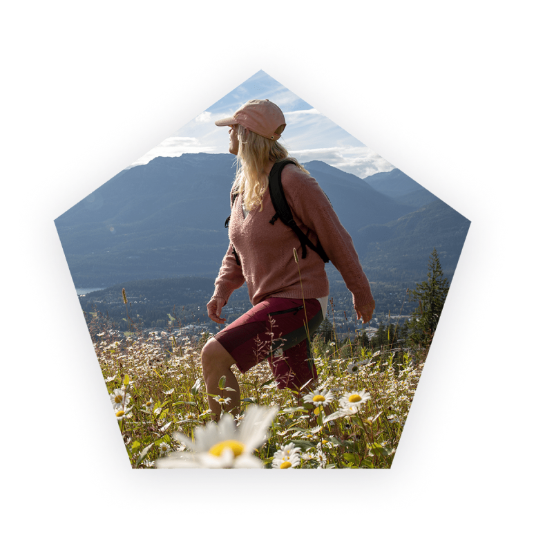 Woman wearing a hat hiking through a field of flowers and mountains in the background.