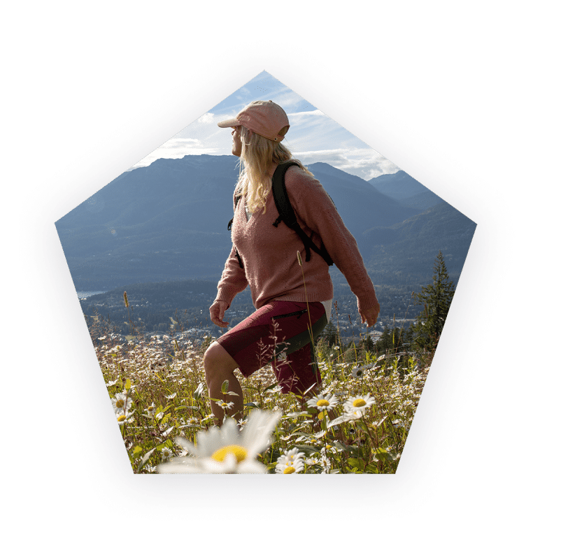 Woman wearing a hat hiking through a field of flowers and mountains in the background.