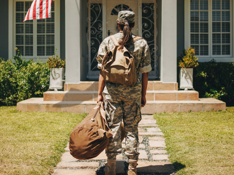 Soldier coming home, carrying a bag and looking at a porch with an American flag.